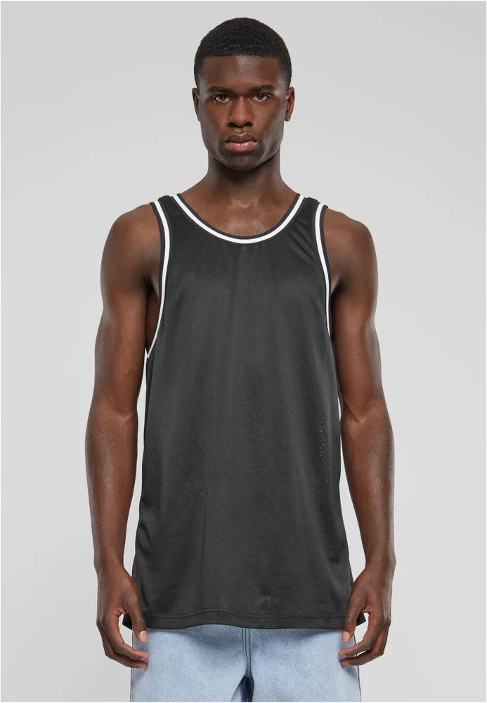 Das "Mesh Tanktop" ist ein schwarzes, ärmelloses Tanktop mit weißen Rändern und einem sportlichen, luftigen Mesh-Stoff. Der Mann trägt das Tanktop zusammen mit blauen Jeans und sieht direkt in die Kamera. Der Hintergrund ist neutral und weiß.